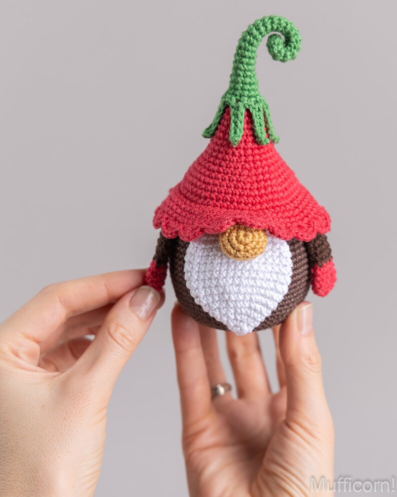 May include: A crocheted gnome ornament with a red hat, brown body, white beard, and a green curly hat.