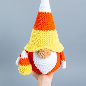 May include: A crocheted candy corn gnome with a white, orange, and yellow hat. The gnome is holding a smaller candy corn in its hand.