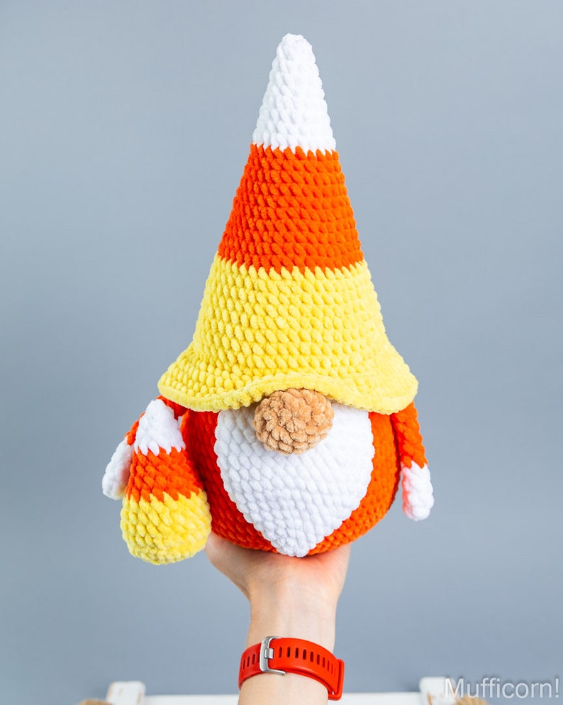 May include: A crocheted candy corn gnome with a white, orange, and yellow hat. The gnome is holding a smaller candy corn in its hand.