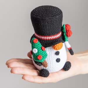 May include: A crocheted snowman holding a small green Christmas tree. The snowman is wearing a black top hat with a red and white stripe, and a red berry wreath.