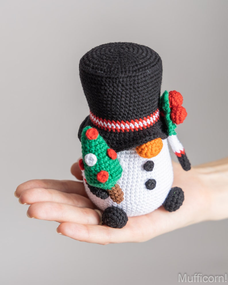 May include: A crocheted snowman holding a small green Christmas tree. The snowman is wearing a black top hat with a red and white stripe, and a red berry wreath.
