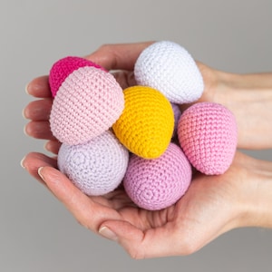 May include: A collection of crocheted Easter eggs in various pastel colors, including pink, purple, yellow, and white. The eggs are held in a person's hands.
