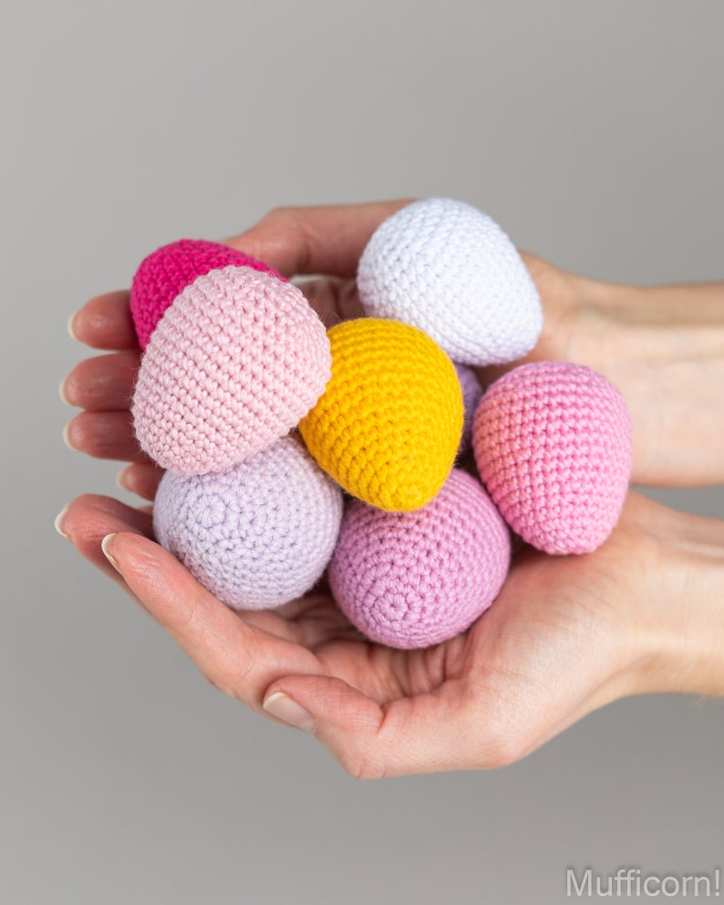 May include: A collection of crocheted Easter eggs in various pastel colors, including pink, purple, yellow, and white. The eggs are held in a person's hands.