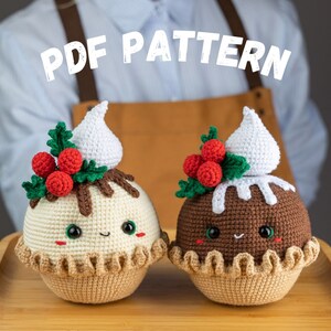May include: Two crocheted Christmas pudding ornaments with smiling faces, red holly berries, and white frosting. The ornaments are on a wooden surface. PDF PATTERN