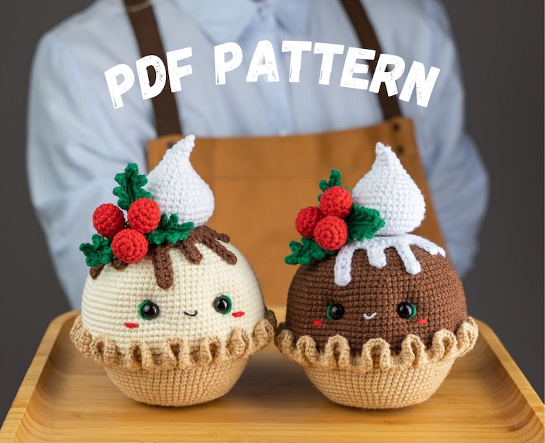 May include: Two crocheted Christmas pudding ornaments with smiling faces, red holly berries, and white frosting. The ornaments are on a wooden surface. PDF PATTERN
