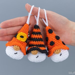 May include: Three handmade crochet gnome ornaments with white bodies, tan beards, and smiling faces. Each gnome has a different orange and black hat design, with white hanging loops. Perfect for Halloween or fall decor.