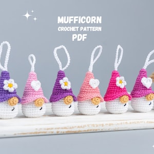 May include: A collection of six crocheted gnome ornaments in various shades of purple, pink, and white. Each gnome has a conical hat, a white beard, and a small flower or heart detail. The text "MUFFICORN CROCHET PATTERN PDF" is displayed above the gnomes.