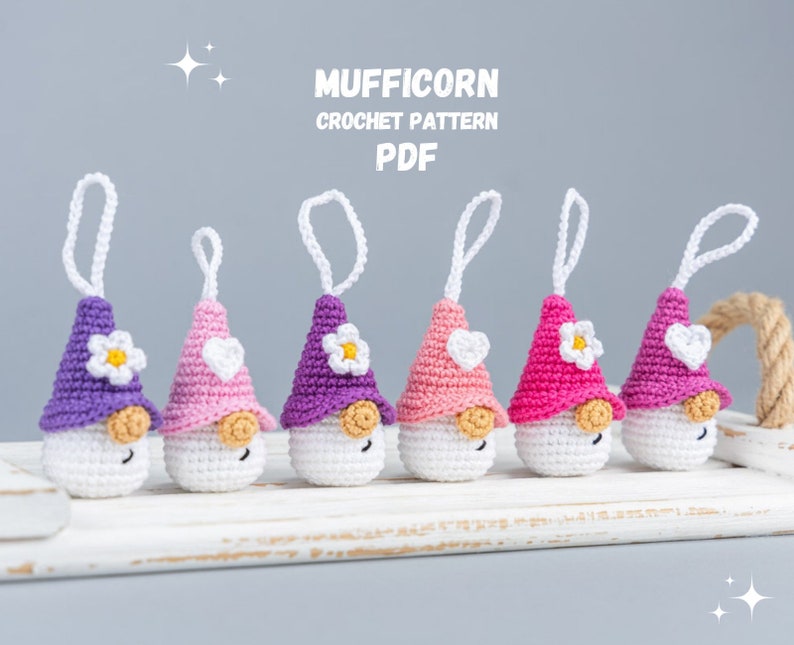 May include: A collection of six crocheted gnome ornaments in various shades of purple, pink, and white. Each gnome has a conical hat, a white beard, and a small flower or heart detail. The text "MUFFICORN CROCHET PATTERN PDF" is displayed above the gnomes.