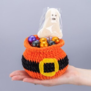 May include: A small orange crochet pouch with a black belt and yellow buckle design, filled with several round chocolate candies wrapped in purple, black, and orange foil, and a single white ghost-shaped lollipop in a clear plastic wrapper.