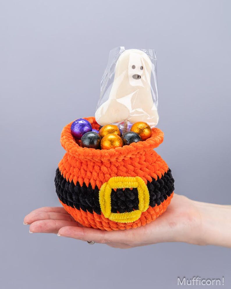 May include: A small orange crochet pouch with a black belt and yellow buckle design, filled with several round chocolate candies wrapped in purple, black, and orange foil, and a single white ghost-shaped lollipop in a clear plastic wrapper.