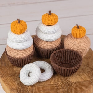 May include: Three crocheted pumpkin cupcakes and a donut on a wooden cutting board. The cupcakes are stacked with white and brown layers and topped with orange pumpkins. The donut is white and has a hole in the center.