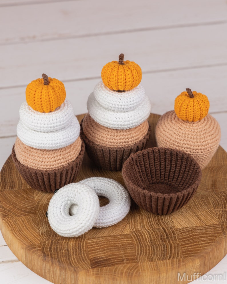 May include: Three crocheted pumpkin cupcakes and a donut on a wooden cutting board. The cupcakes are stacked with white and brown layers and topped with orange pumpkins. The donut is white and has a hole in the center.