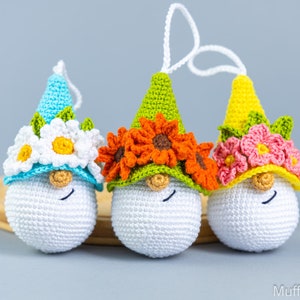 May include: Three crocheted gnome ornaments with white faces and different colored hats. The gnome on the left has a light blue hat with white flowers, the middle gnome has a green hat with orange flowers, and the gnome on the right has a yellow hat with pink flowers.