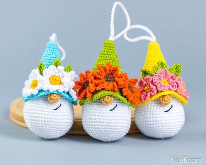 May include: Three crocheted gnome ornaments with white faces and different colored hats. The gnome on the left has a light blue hat with white flowers, the middle gnome has a green hat with orange flowers, and the gnome on the right has a yellow hat with pink flowers.