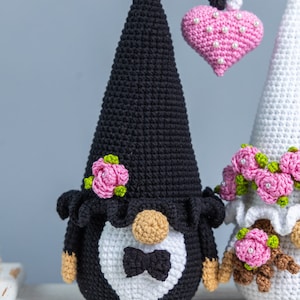 May include: A pair of crocheted gnomes dressed as a bride and groom. The groom gnome is wearing a black hat and a white bow tie. The bride gnome is wearing a white dress and a pink flower crown. They are holding a pink heart.