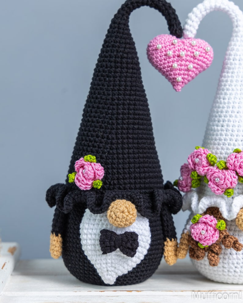 May include: A pair of crocheted gnomes dressed as a bride and groom. The groom gnome is wearing a black hat and a white bow tie. The bride gnome is wearing a white dress and a pink flower crown. They are holding a pink heart.