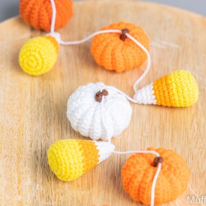 May include: A garland of crocheted pumpkins and candy corn, strung together with white yarn. The garland is displayed on a wooden surface.