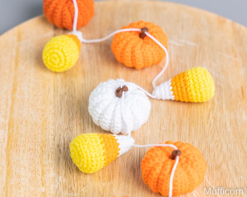 May include: A garland of crocheted pumpkins and candy corn, strung together with white yarn. The garland is displayed on a wooden surface.