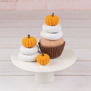 May include: A white cake stand with a brown and tan crocheted cupcake topped with a small orange pumpkin. Two smaller orange pumpkins are stacked on top of two white crocheted circles.