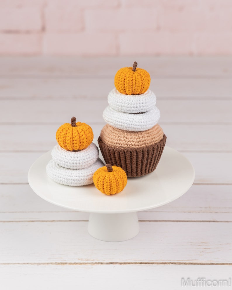 May include: A white cake stand with a brown and tan crocheted cupcake topped with a small orange pumpkin. Two smaller orange pumpkins are stacked on top of two white crocheted circles.