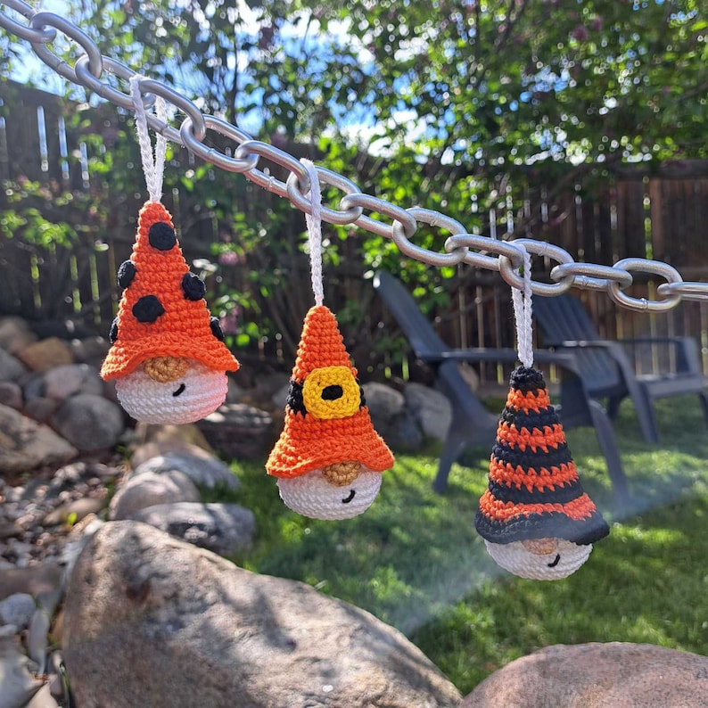 May include: Three crochet gnome ornaments hanging from a silver chain. Each gnome has a white face, a beige beard, and a colorful hat. The hats are orange with black spots, orange with a yellow buckle, and black and orange stripes.