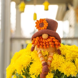 May include: A crocheted gnome wearing a brown hat with a yellow flower on top. The gnome is sitting in a pot of yellow chrysanthemums.