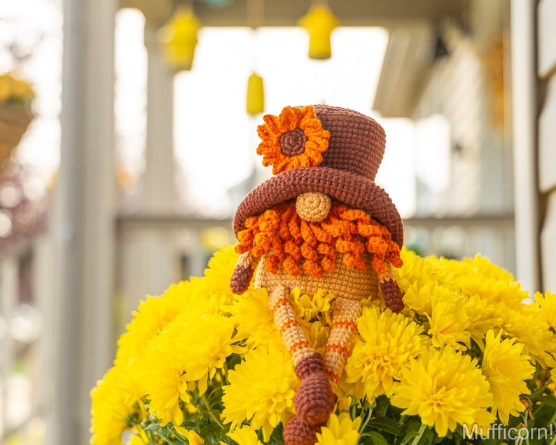 May include: A crocheted gnome wearing a brown hat with a yellow flower on top. The gnome is sitting in a pot of yellow chrysanthemums.