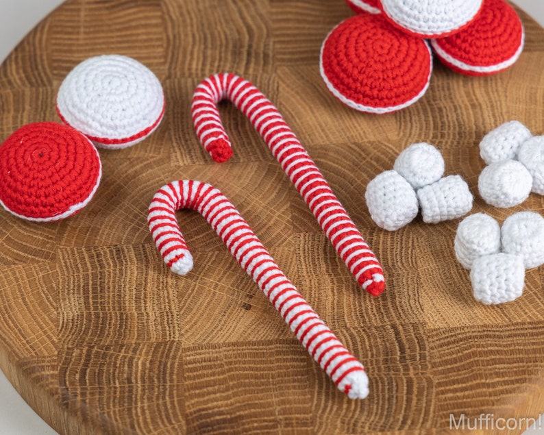 Puede incluir: Art&iacute;culos navide&ntilde;os de ganchillo, que incluyen bastones de caramelo a rayas rojas y blancas, adornos redondos rojos y blancos y formas de malvavisco de ganchillo blanco. Los art&iacute;culos est&aacute;n dispuestos sobre una superficie de madera.