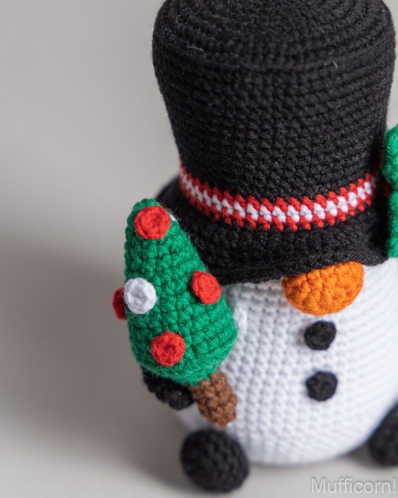 May include: A crocheted snowman holding a small green Christmas tree with red and white ornaments. The snowman is wearing a black top hat with a red and white band.