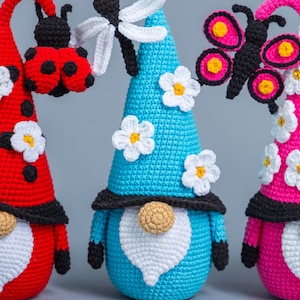 May include: Three crocheted gnomes with different colored hats and insect decorations. The gnome on the left has a red hat with a ladybug, the middle gnome has a blue hat with a dragonfly, and the gnome on the right has a pink hat with a butterfly.