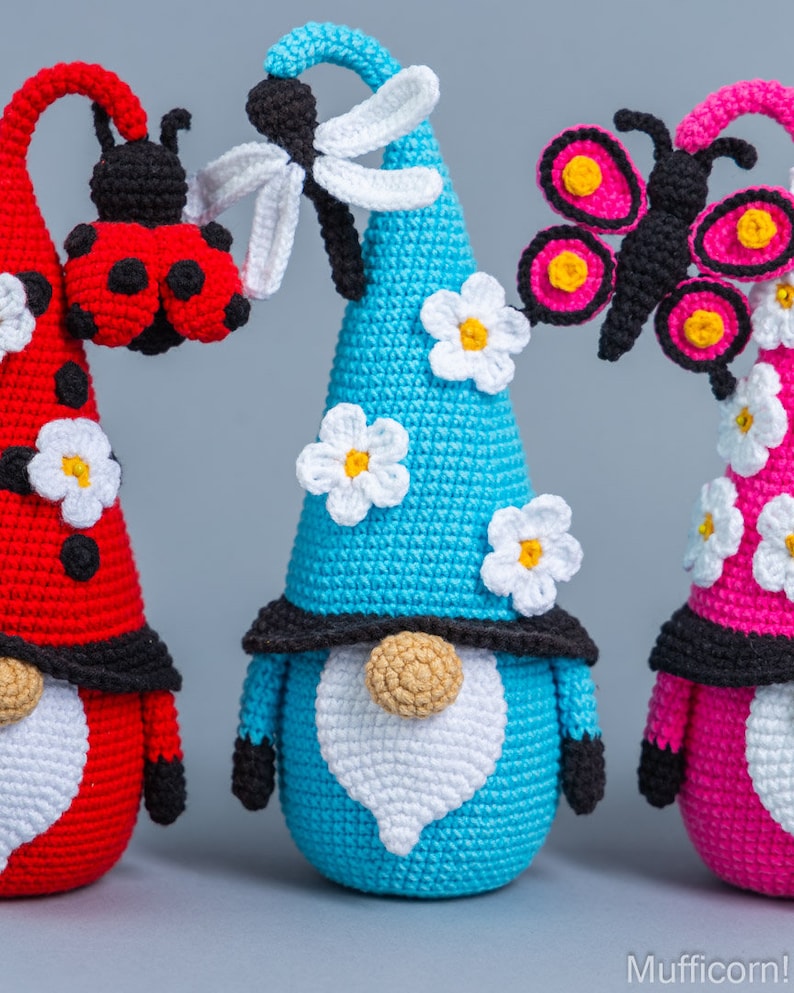 May include: Three crocheted gnomes with different colored hats and insect decorations. The gnome on the left has a red hat with a ladybug, the middle gnome has a blue hat with a dragonfly, and the gnome on the right has a pink hat with a butterfly.