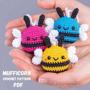 Puede incluir: Tres adornos de abeja de ganchillo en rosa, azul y amarillo, cada uno con rayas negras, antenas y caras sonrientes. Alas blancas están unidas. El texto "MUFFICORN CROCHET PATTERN PDF" es visible en la parte inferior.