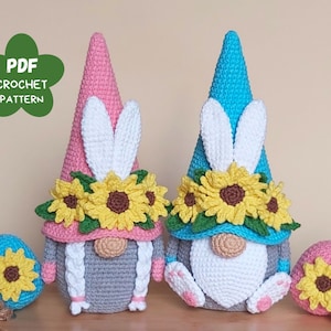 May include: Two crocheted Easter gnomes with sunflower crowns and bunny ears. One gnome is wearing a pink hat and the other is wearing a blue hat. Each gnome is holding a crocheted Easter egg.
