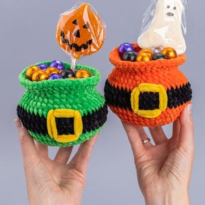 May include: Two crocheted Halloween treat baskets, one green and one orange, each featuring a black belt with a yellow buckle. The green basket holds a pumpkin lollipop, and the orange basket holds a ghost lollipop, both filled with colorful candies.