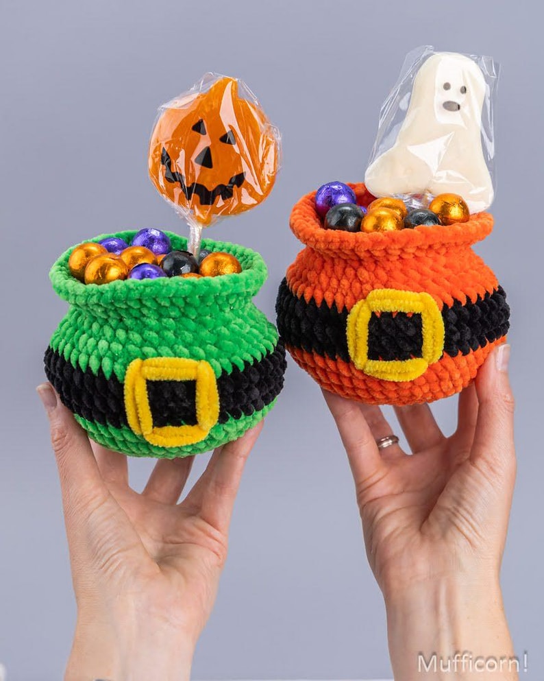 May include: Two crocheted Halloween treat baskets, one green and one orange, each featuring a black belt with a yellow buckle. The green basket holds a pumpkin lollipop, and the orange basket holds a ghost lollipop, both filled with colorful candies.