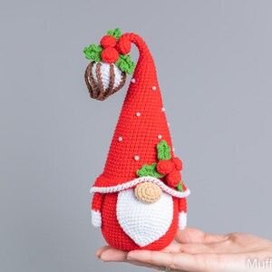 May include: A red crocheted gnome with a white beard and a red hat with white dots and green holly. The gnome is holding a brown and white crocheted ornament.