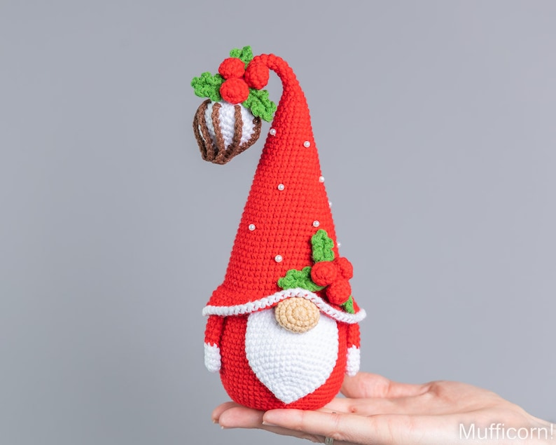 May include: A red crocheted gnome with a white beard and a red hat with white dots and green holly. The gnome is holding a brown and white crocheted ornament.