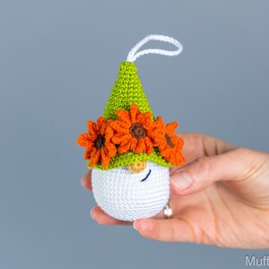 May include: A white crocheted gnome ornament with a green hat and orange flower accents. The gnome has a small, white beard and a closed eye.