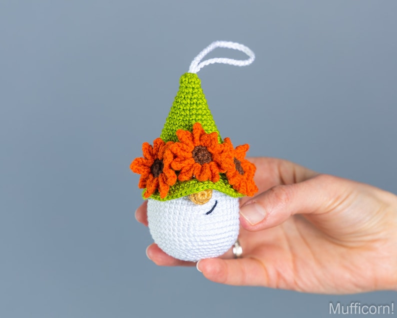 May include: A white crocheted gnome ornament with a green hat and orange flower accents. The gnome has a small, white beard and a closed eye.