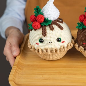 May include: Two crocheted Christmas pudding ornaments on a wooden tray. The pudding on the left has a white crocheted whipped cream topping, green holly leaves, and red berries. The pudding on the right is brown with a green holly leaf and red berries.