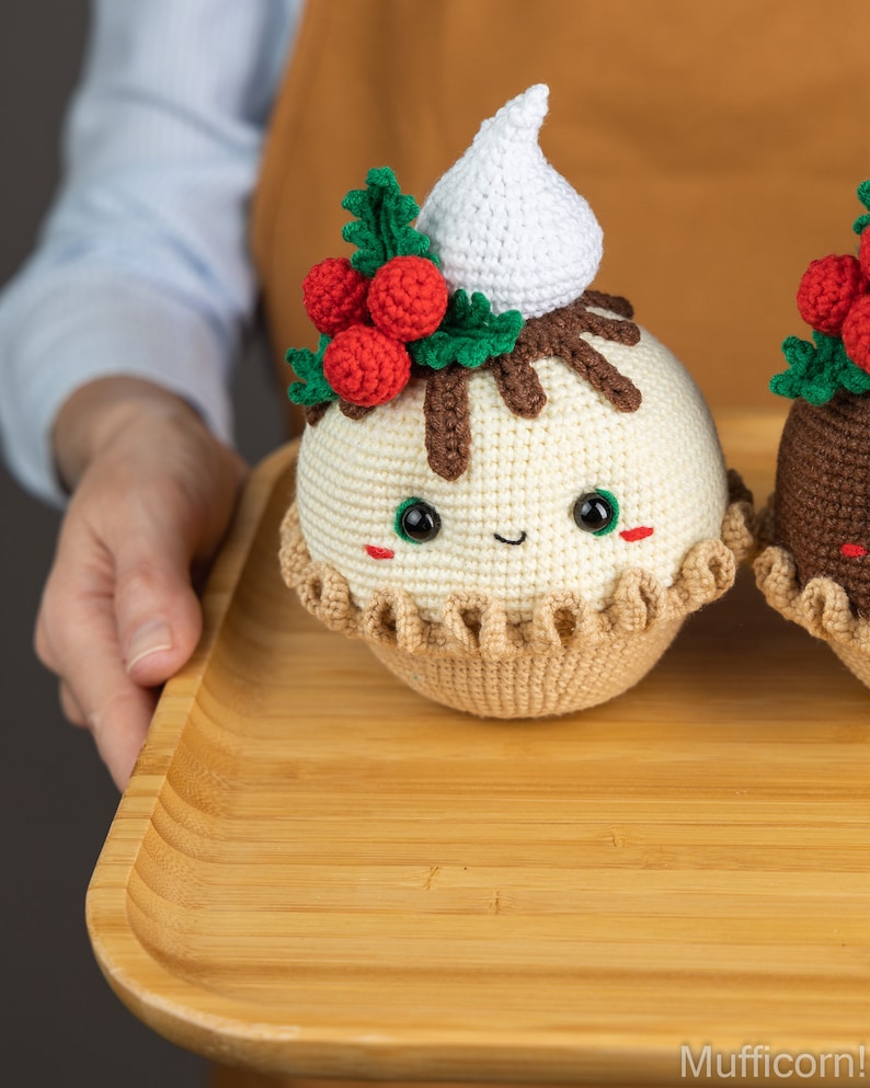 May include: Two crocheted Christmas pudding ornaments on a wooden tray. The pudding on the left has a white crocheted whipped cream topping, green holly leaves, and red berries. The pudding on the right is brown with a green holly leaf and red berries.