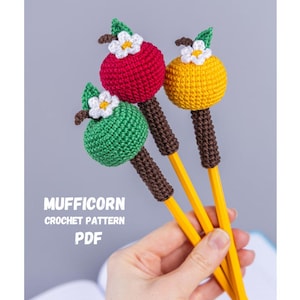 Peut inclure: Trois crayons avec des dessus en crochet en forme de pommes rouges, vertes et jaunes. Chaque pomme a une feuille verte et une fleur blanche. Les crayons ont des tiges marron et sont tenus dans une main. Le texte "MUFFICORN CROCHET PATTERN PDF" est visible.