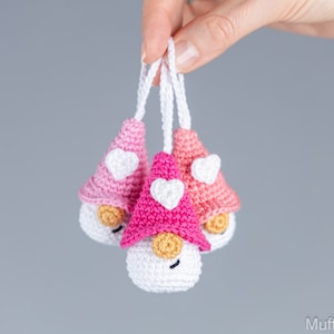May include: Three handmade crochet gnomes with white bodies, tan noses, and smiling faces. Each gnome has a different shade of pink hat with a white heart. The gnomes are suspended by white strings.