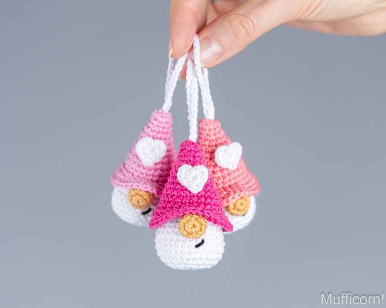 May include: Three handmade crochet gnomes with white bodies, tan noses, and smiling faces. Each gnome has a different shade of pink hat with a white heart. The gnomes are suspended by white strings.