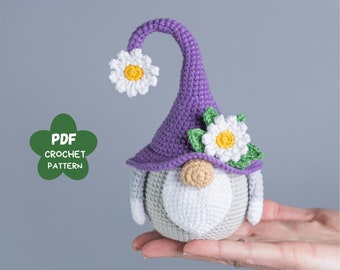 Op de afbeelding: Een gehaakte gnome met een paarse hoed en witte bloemen. De gnome heeft een witte baard en een bruine neus. De gnome houdt een bloem in zijn hand.