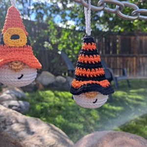 May include: Two handmade crochet gnome ornaments. Each gnome has a white body, a pointed hat in orange and black, and a small, smiling face. The ornaments are hanging from a silver chain in an outdoor setting.