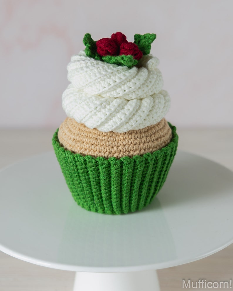 May include: A crocheted cupcake with green icing and a white swirl on top. The cupcake is decorated with red berries and green leaves.