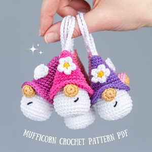 May include: Three handmade crochet gnomes with white bodies, tan noses, and smiling faces. Each gnome wears a different colored hat: pink, purple, and pink. The hats are adorned with white flowers. The text "MUFFICORN CROCHET PATTERN PDF" is below the gnomes.
