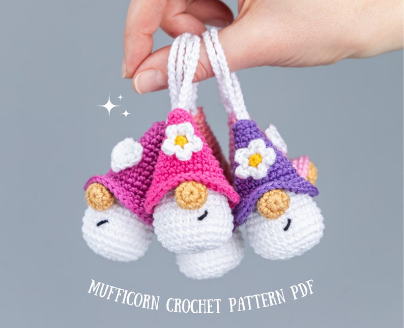 May include: Three handmade crochet gnomes with white bodies, tan noses, and smiling faces. Each gnome wears a different colored hat: pink, purple, and pink. The hats are adorned with white flowers. The text "MUFFICORN CROCHET PATTERN PDF" is below the gnomes.