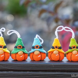 May include: Five crocheted pumpkin gnomes with different colored hats. The gnomes are wearing black belts and have orange pumpkin bodies. The gnomes are hanging from white strings.